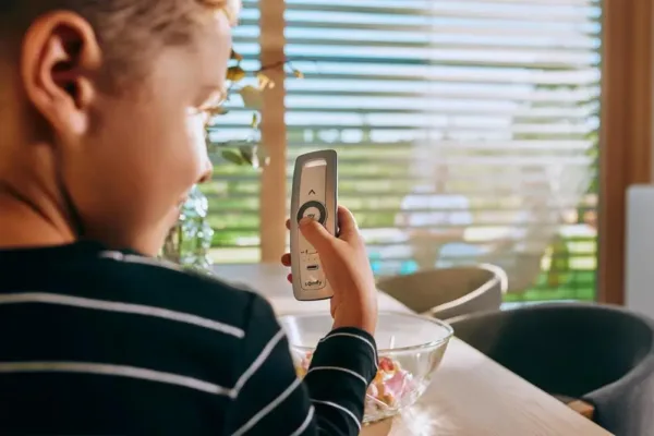 Ein Kind hält eine Fernbedienung in der Nähe eines Fensters, durch dessen Jalousien Sonnenlicht fällt.