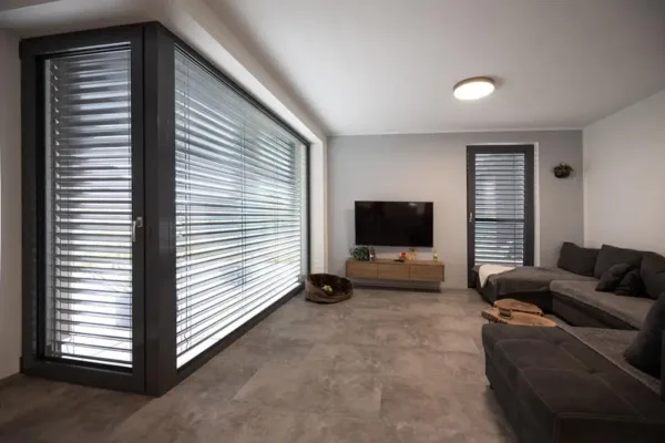 Modernes Wohnzimmer mit grauen Sofas, TV, großen Fenstern mit Jalousien und minimalem Dekor.