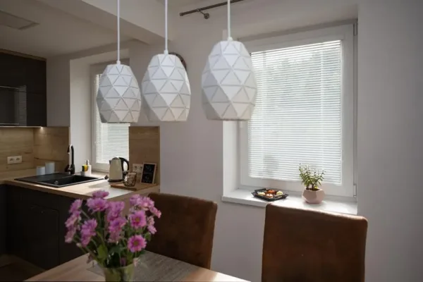 Moderne Küche mit Hängeleuchten, Blumen auf dem Tisch und Sonnenlicht durch die Jalousien.