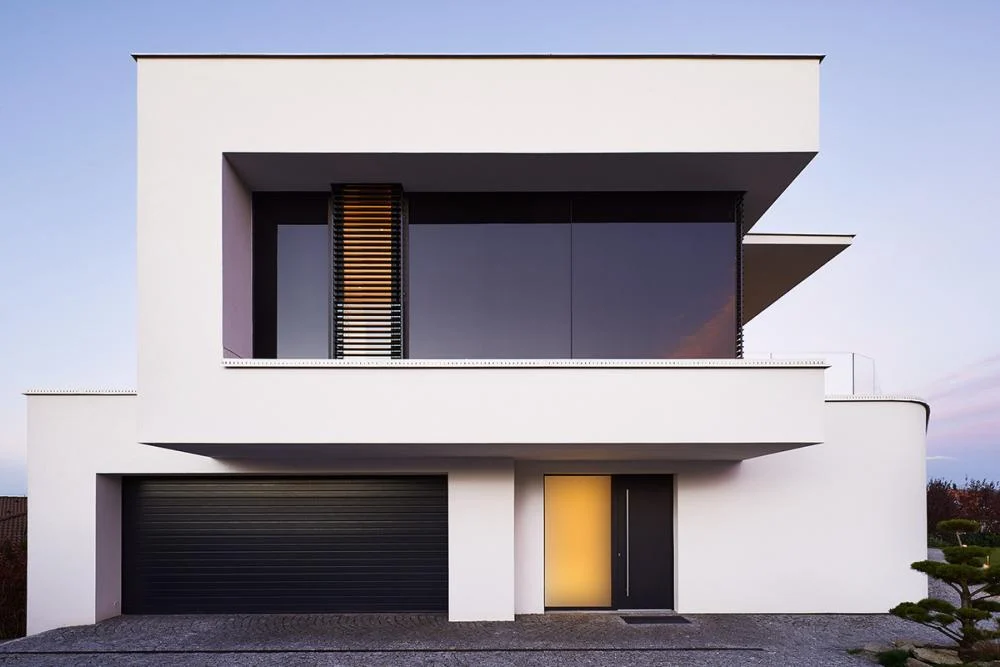 Modernes weißes zweistöckiges Haus mit großen Fenstern, einer schwarzen Garage und einer leuchtenden Eingangstür.