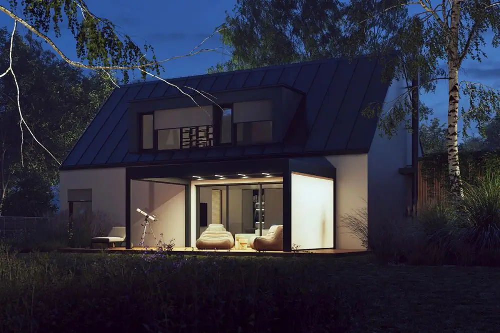 Modernes Haus mit großen Fenstern und Patio, warm beleuchtet an einem späteren Abend, umgeben von Bäumen.