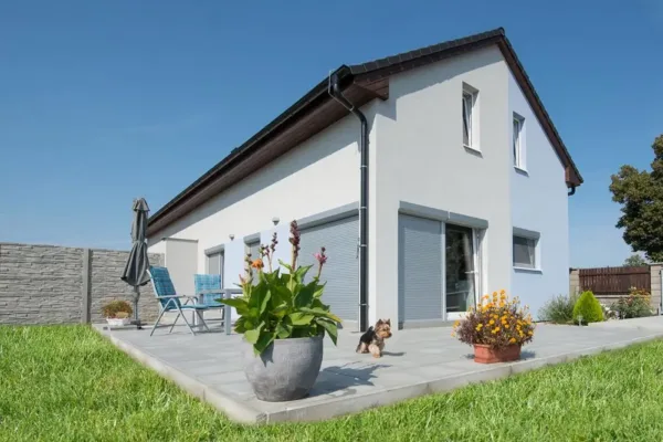Modernes weißes Haus mit Terrasse, Topfpflanzen, Sitzgelegenheiten im Freien und einem kleinen Hund auf der Wiese.