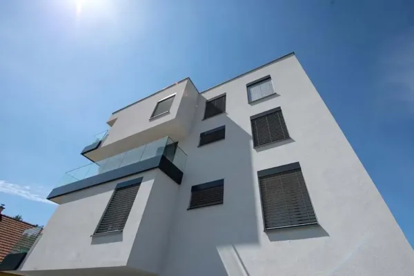 Modernes weißes Mehrfamilienhaus mit Balkonen und geschlossenen Fensterläden unter einem klaren blauen Himmel.